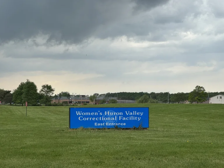 A sign for Women’s Huron Valley Correctional Facility stands in a grassy area with the prison visible in the distance under dark storm clouds.