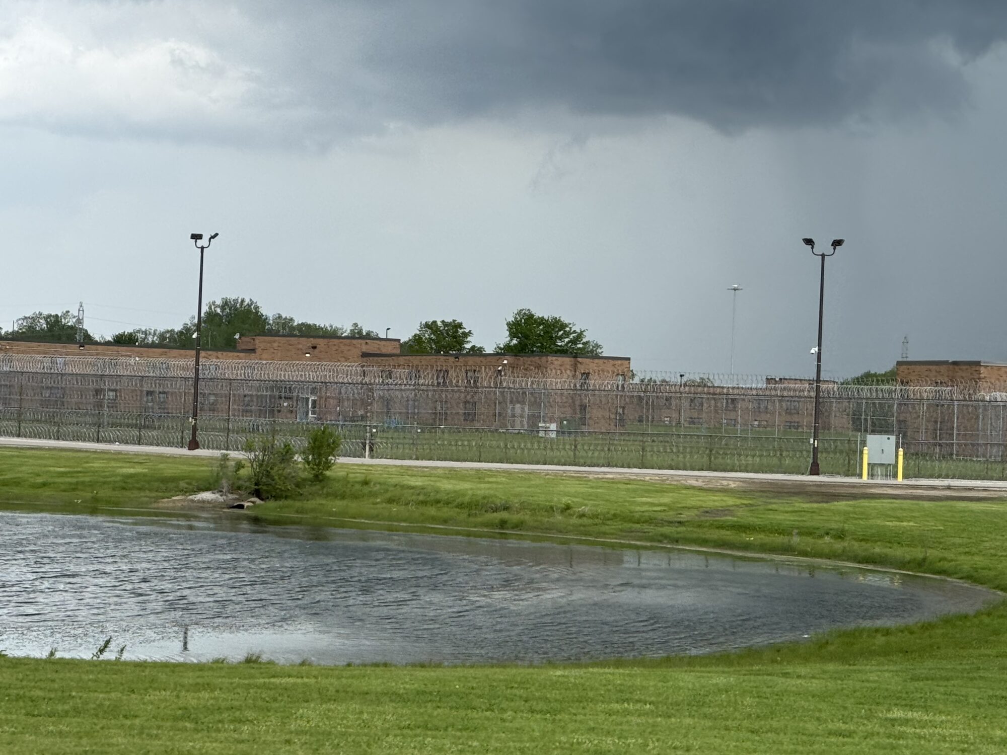 A view of Women’s Huron Valley Correctional Facility behind a security fence, with grass, water and dark clouds in the foreground.
