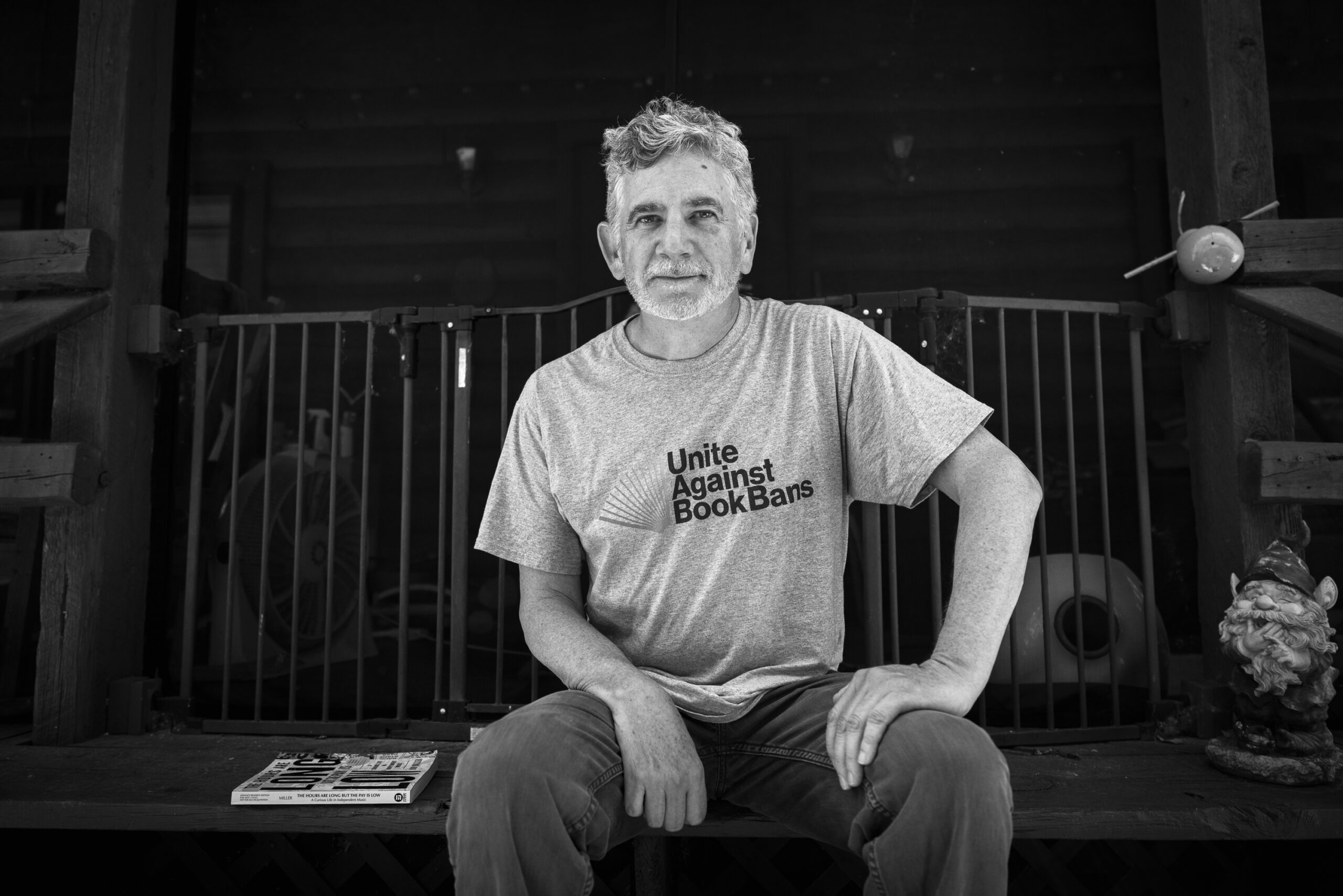 Rob Miller sits on a wooden porch beside a copy of his book, wearing a “Unite Against Book Bans” T-shirt.